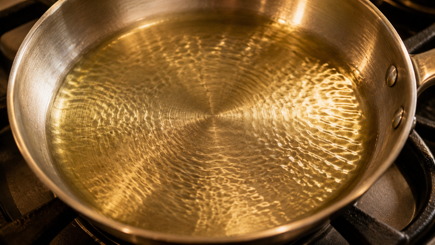 A stainless steel pan with a thin layer of shimmering cooking oil, showing visible ripples across the surface in warm kitchen light.